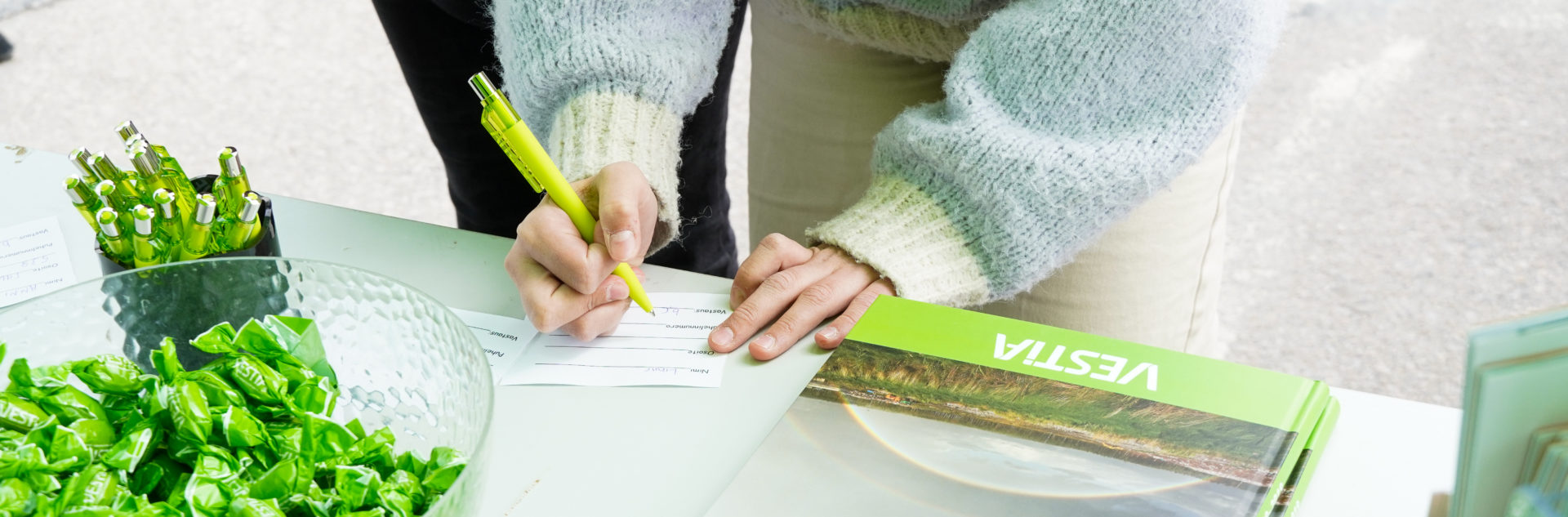 Kädet kirjoittamassa arvontalappua pöydällä, jossa Vestian kyniä, karkkeja ja kirjoja. Kädet kirjoittamassa arvontalappua pöydällä, jossa Vestian kyniä, karkkeja ja kirjoja.