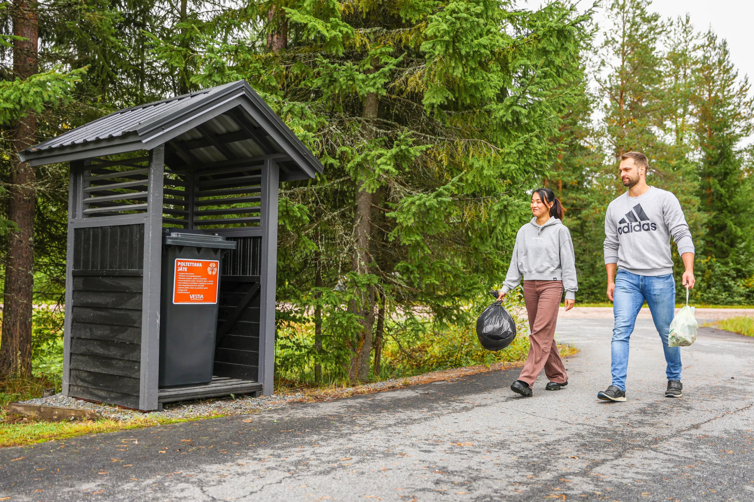 Kaksi henkilöä lähestyvät jäteastiaa roskapussit kädessä