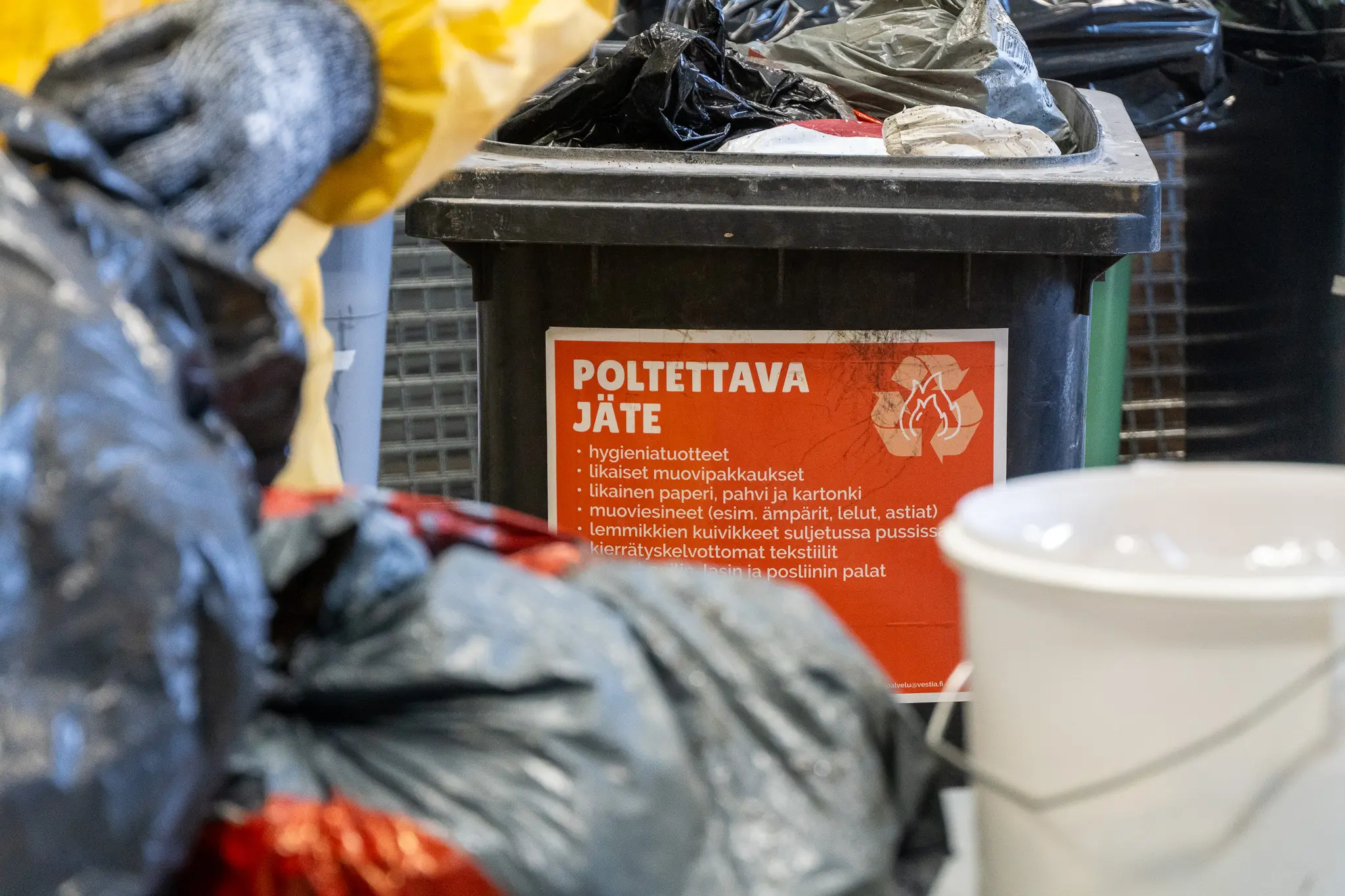 Kuvassa poltettavan jätteen astia, jossa roskapusseja. Kuvan etualalla näkyy lisää roskapusseja, valkoinen ämpäri ja suojavarusteisiin pukeutuneen henkilön käsi.