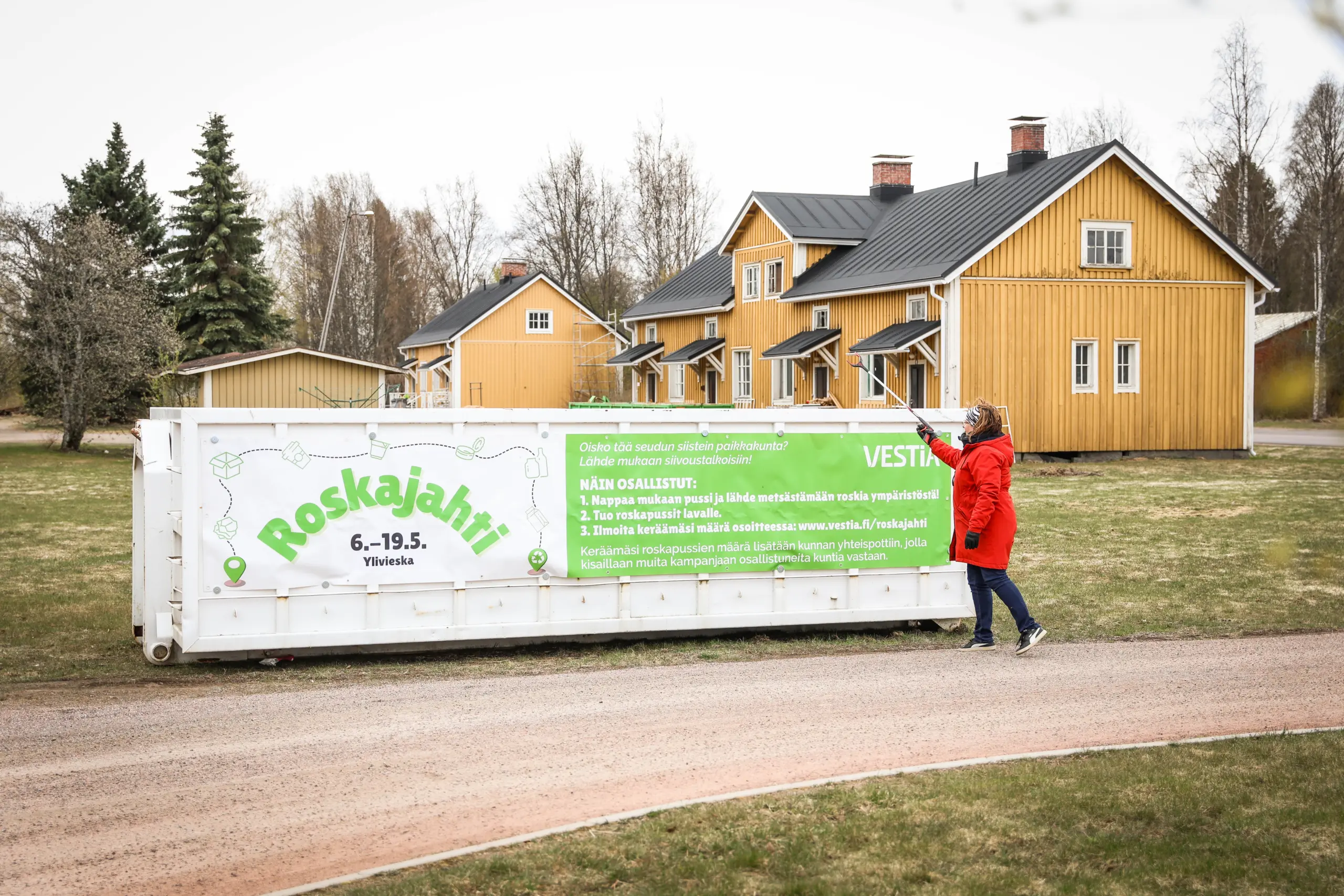 Roskajahti-lava ja henkilö laittamassa roskapihdeillä roskaa lavalle. Taustalla rakennuksia ja puita.