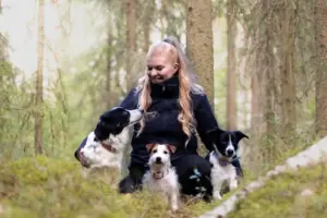 henkilö kyykyssä metsän siimeksessä, ympärillä kolme koiraa.