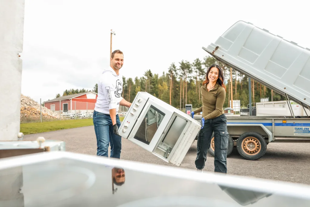 Kaksi henkilöä kantavat rikkinäistä uunia peräkärrystä pois lajittelupihalla