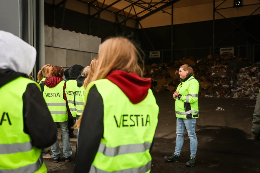 Kierrätysneuvoja ison ryhmän edessä jätehallin edessä kertomassa jätteiden kierrätyksestä, vierailijoilla Vestian heijastinliivit päällä