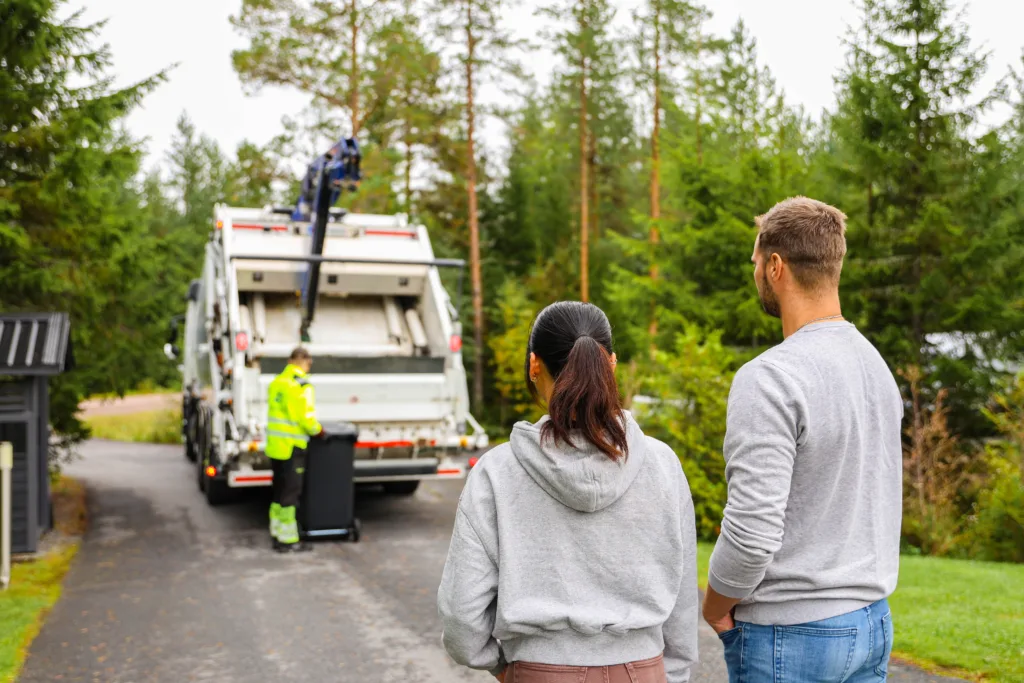Kaksi henkilö etualalla selin katsoo takana näkyvää jäteautoa, johon jätekuski on tyhjentämässä jäteastiaa