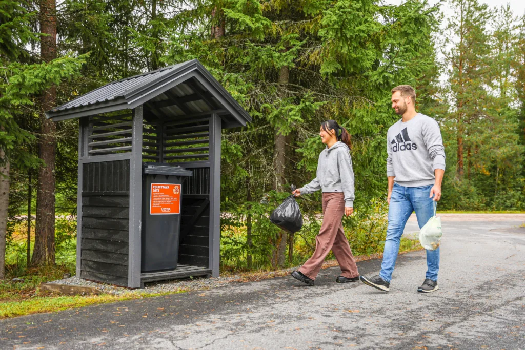Kaksi henkilöä kävelevät omakotitalon pihatietä pitkin kohti roskakatosta roskapussit kädessä